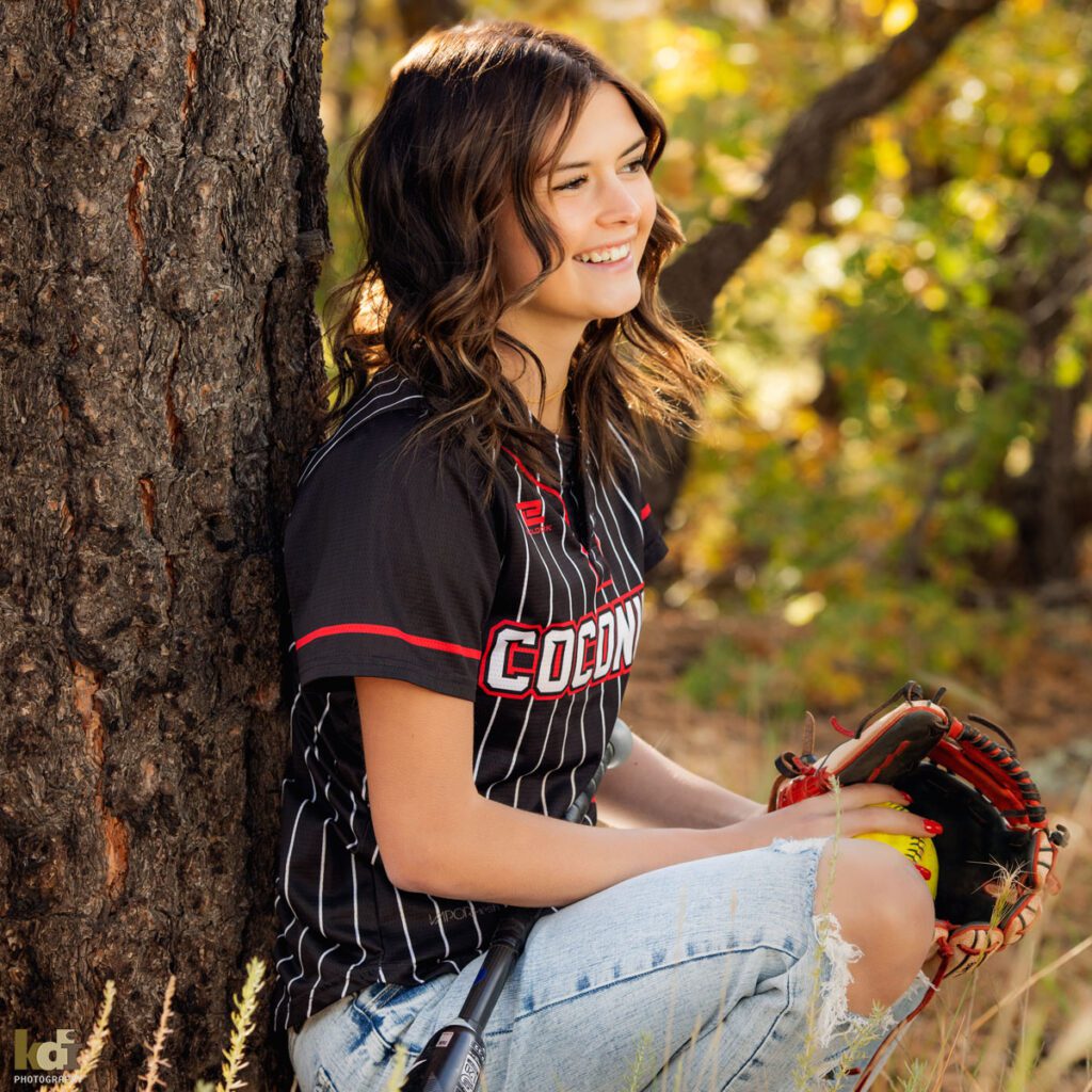 Coconino High School softball player sits against tree, Flagstaff senior photos, by KDI Photography.