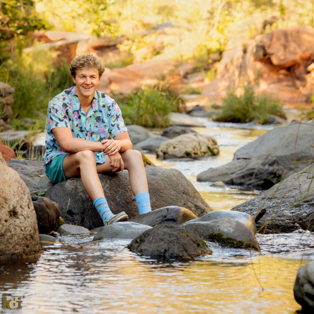 High school senior boy, sitting on a rock in the creek in Hawaiian shirt and blue fish socks, Sedona, AZ. Senior photos by Flagstaff Portrait photographer, KDI Photography.