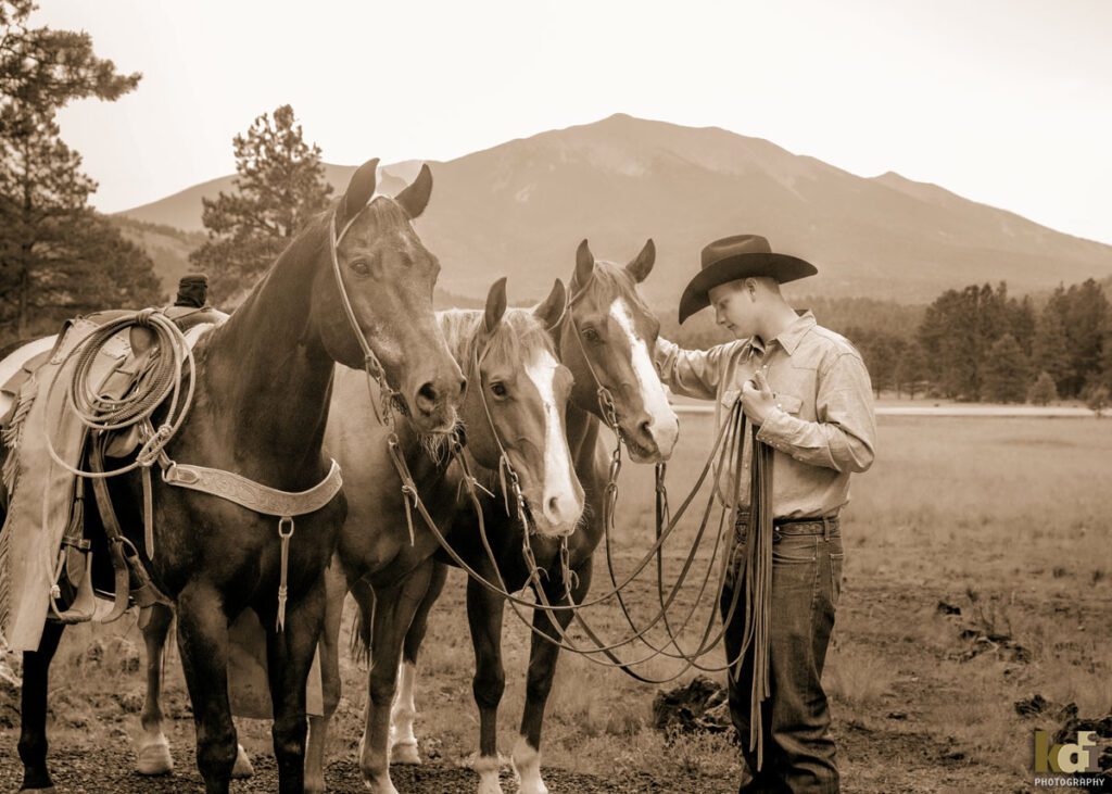 Black and white senior photo of cowboy holding lead ropes with three horses in the forest near Flagstaff, AZ, KDI Photography