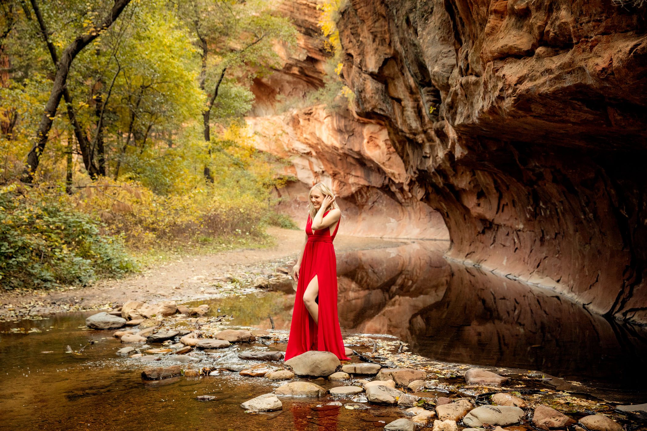 Young Woman in Red Dress, Tucks her Hair Behind Her Ear While Crossing the Creek in a Res Rock Slot Canyon in the Background, Sedona Senior Photos, Fall in Flagstaff, AZ KDI Photography