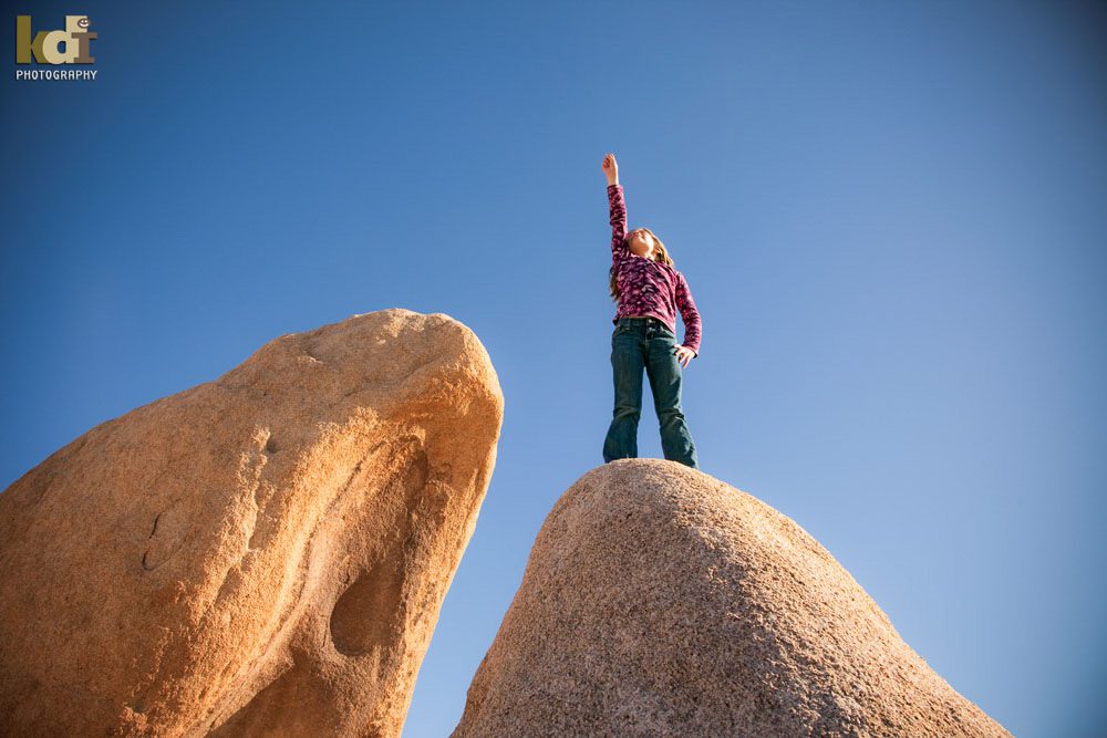 Color location portrait of a tween preteen girl standing pin top of boulders in Joshua Tree National Park, looking up, her fist in the air, family pictures in Northern Arizona, family photographs by KDI Photography