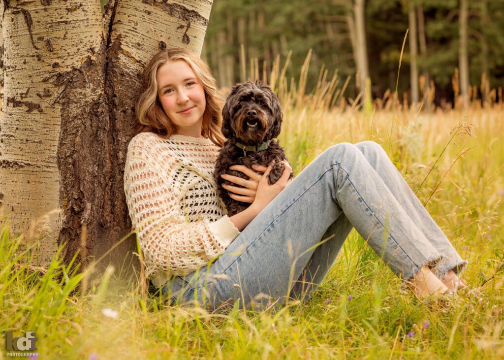 high school senior photo showing girl with reddish light brown hair holding black dog, in off-white sweater and jeans, looking at camera, sitting in mountain grasses, leaning against large aspen trees, portrait in Flagstaff, AZ by senior photographer KDI Photography