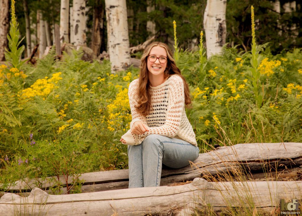 high school senior photo showing girl with reddish light brown hair with glasses, in off-white sweater and jeans, looking at camera laughing while sitting in the mountain ferns and yellow flowers, portrait in Flagstaff, AZ by senior photographer KDI Photography