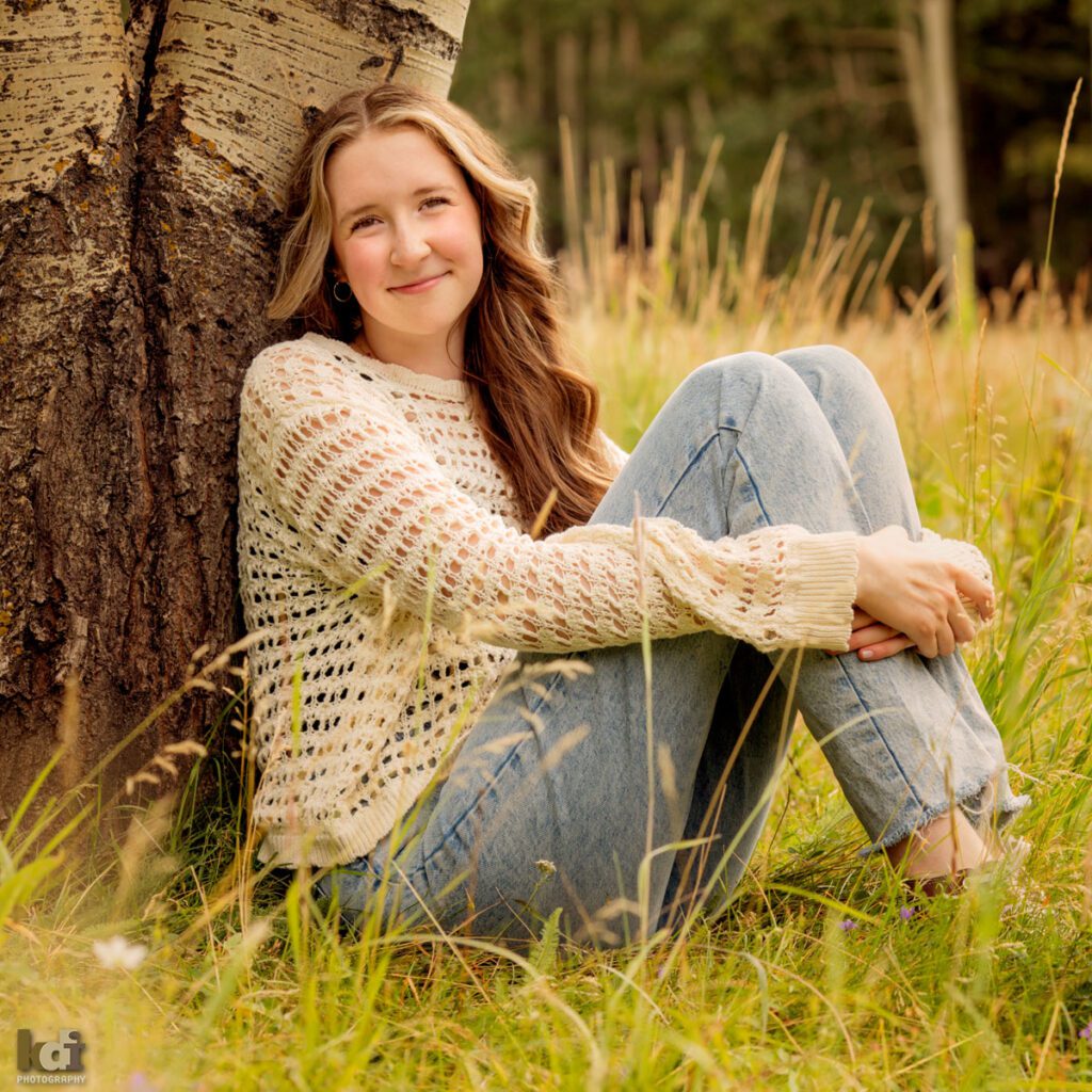 high school senior photo showing girl with reddish light brown hair, in off-white sweater and jeans, looking at camera, sitting in mountain grasses, leaning against large aspen trees, portrait in Flagstaff, AZ by senior photographer KDI Photography