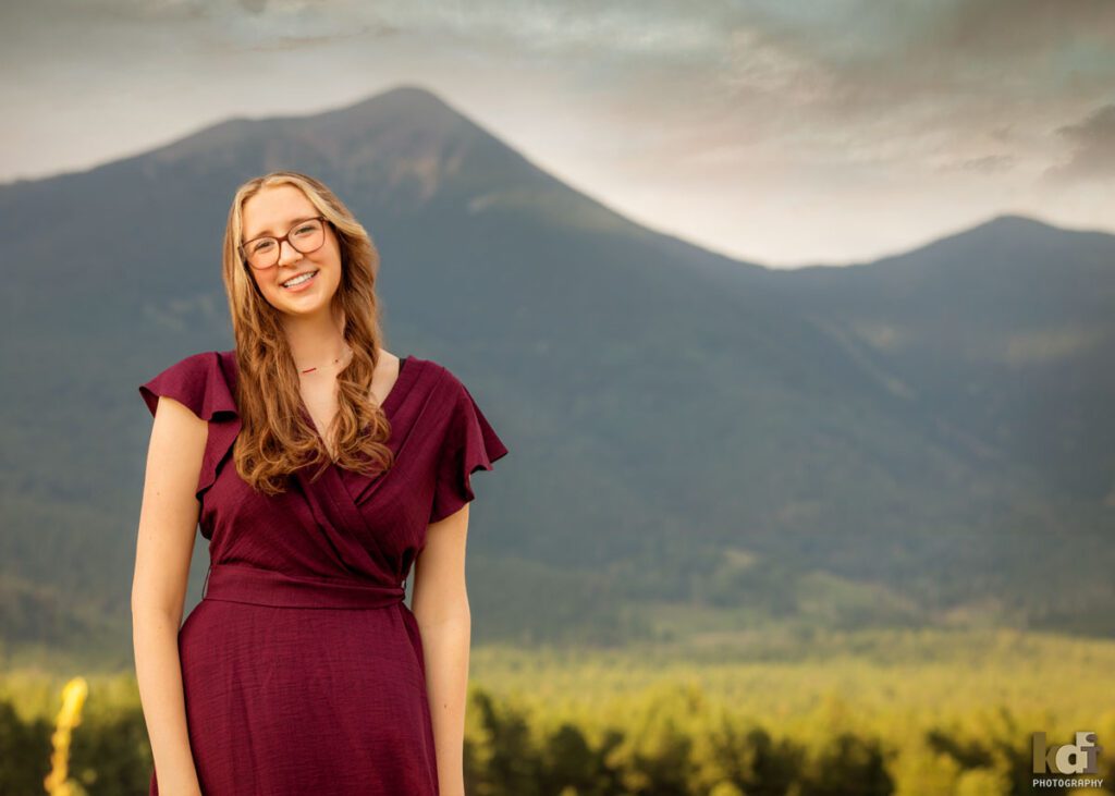 high school senior photo showing girl with reddish light brown hair in purple dress, looking at camera, smiling while standing in front of mountains, San Francisco Peaks, portrait in Flagstaff, AZ by senior photographer KDI Photography