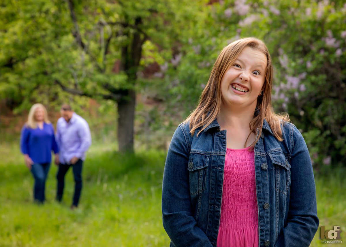 Portrait of mother, father laughing as they stand behind their tween daughter making faces, with lilacs behind them in Sedona AZ, photographic art by KDI Photography. 