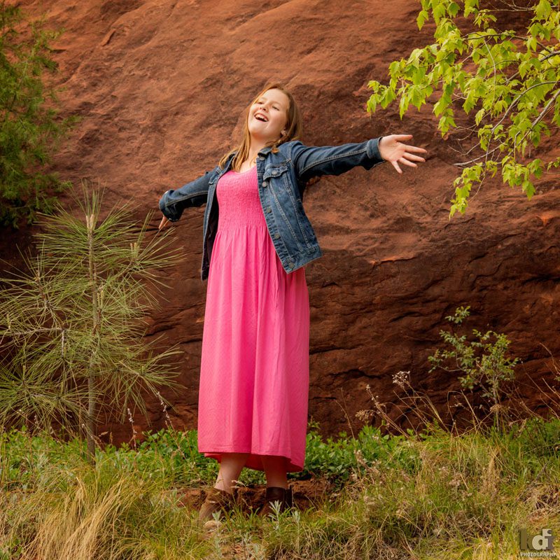 Portrait of tween girl wearing pink dress and jean jacket, standing near red rocks, with arms triumphantly outstretched in Sedona AZ, photographic art by KDI Photography.