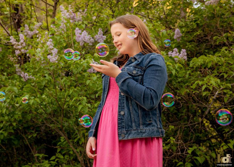 Color portrait of tween girl smiling, looking at and catching bubbles with lilacs behind her, in Sedona AZ, photographic art by KDI Photography