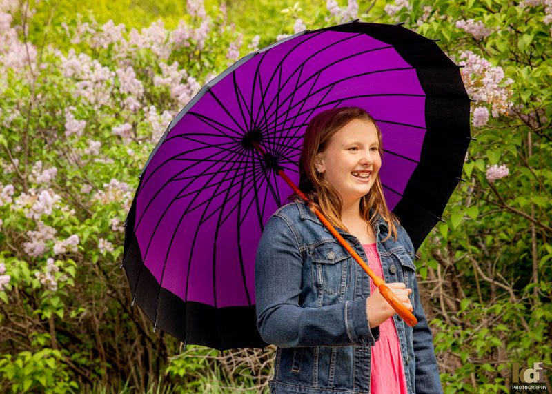 Portrait of tween girl smiling, looking away from camera, holding a purple umbrella with lilacs behind her, in Sedona AZ, photographic art by KDI Photography.