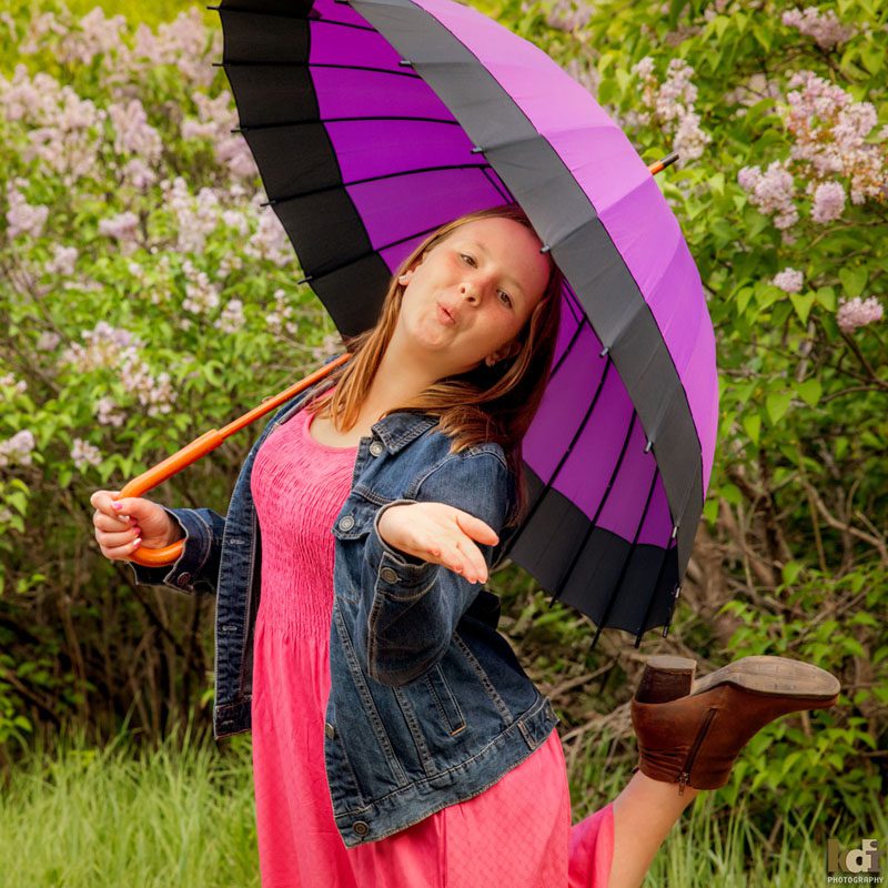 Portrait of tween girl blowing kisses, with her foot kicked up behind her, holding a purple umbrella with lilacs in the background in Sedona AZ, photographic art by KDI Photography.