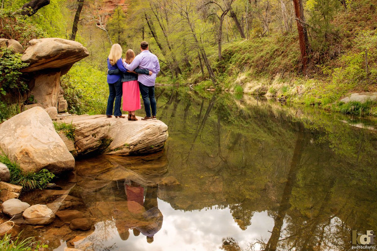 KDI Photography portrait of mother, father and tween daughter standing on rocks with arms around each other by a creek in Sedona AZ, photographic portrait art