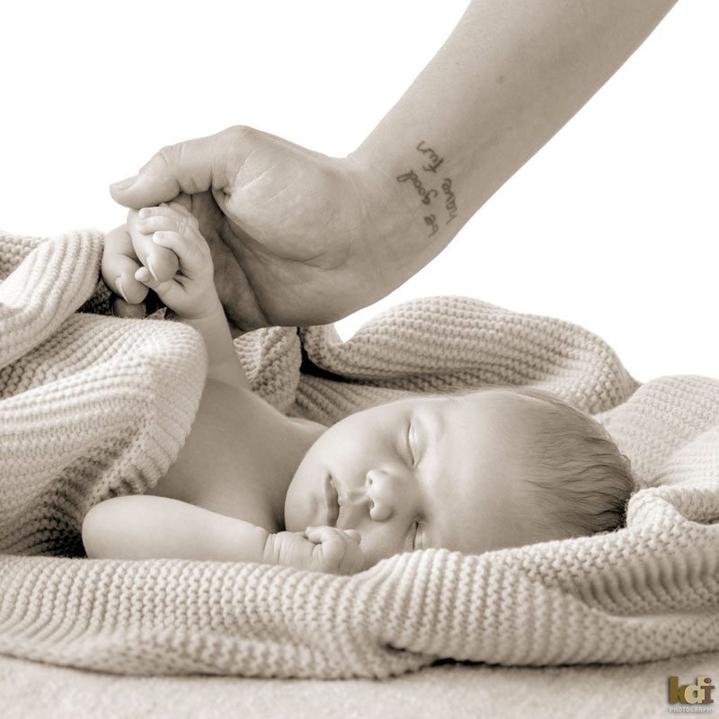 Black and white family photo of a mother's hand holding her newborn girl's hand with a tattoo on her wrist that says - be good have fun - while the baby is calmly asleep, studio portraits in Northern Arizona, family portraits by KDI Photography