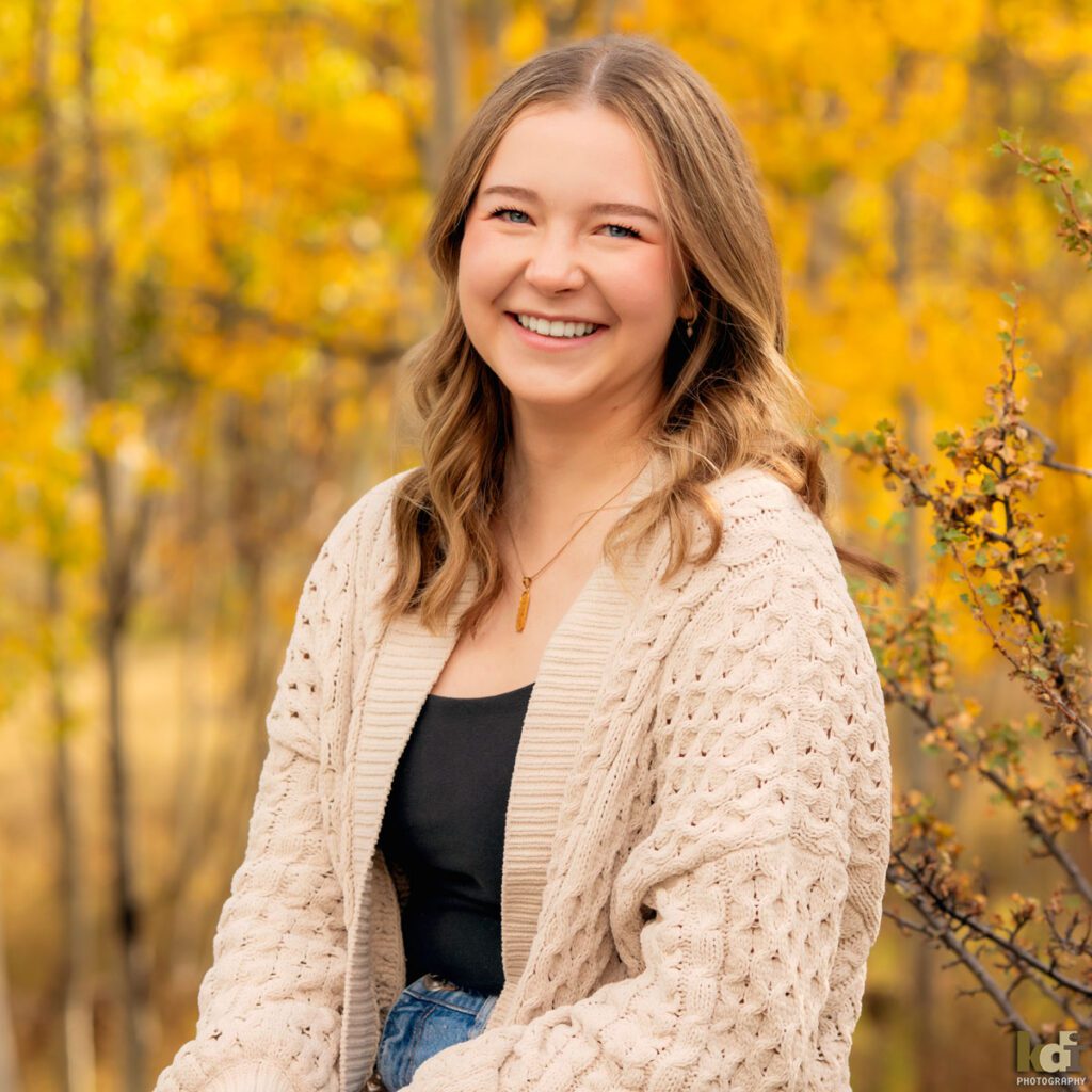 A color photograph of a high school senior photo showing girl with blond hair, in jeans and a white cable knit sweater, laughing at the camera, among the colorful fall aspen trees, portrait photography in Flagstaff AZ by senior photographer KDI Photography