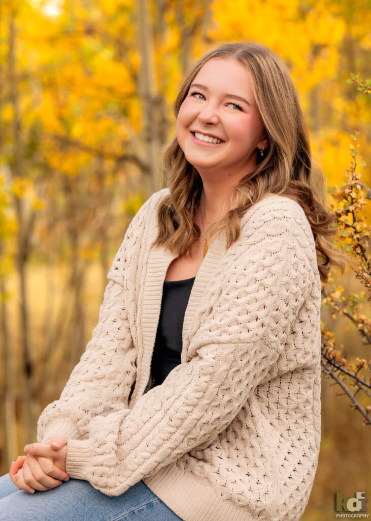 A color photograph of a high school senior photo showing girl with blond hair, in jeans and a white cable knit sweater, laughing, sitting among the colorful fall aspen trees, portrait photography in Flagstaff AZ by senior photographer KDI Photography