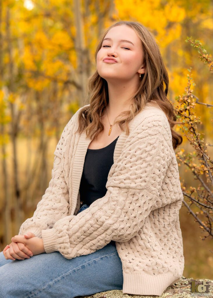 A color photograph of a high school senior photo showing girl with blond hair, in jeans and a white cable knit sweater, making a funny face while sitting among the colorful fall aspen trees, portrait photography in Flagstaff AZ by senior photographer KDI Photography