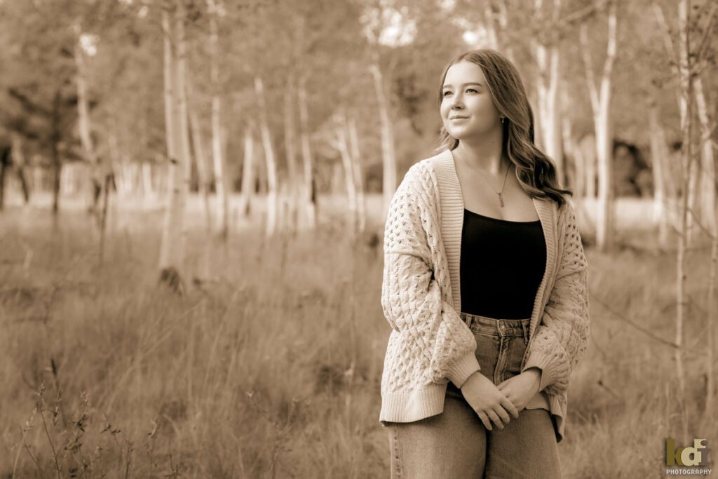 A black and white photograph of a high school senior photo showing girl with blond hair, in jeans and a white cable knit sweater, among the fall aspen trees, portrait photography in Flagstaff AZ by senior photographer KDI Photography