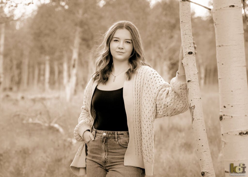 A black and white photograph of a high school senior photo showing girl with blond hair, in jeans and a white cable knit sweater, looking at camera wiht serious expression, among the fall aspen trees, portrait photography in Flagstaff AZ by senior photographer KDI Photography
