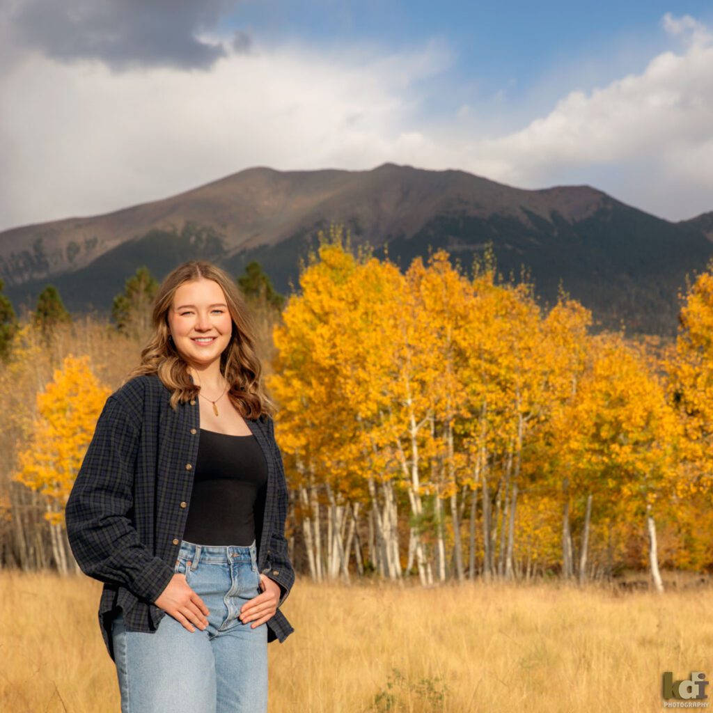 A color photograph of a high school senior photo showing girl with blond hair, in jeans among the fall aspen trees, with the San Francisco Peaks in background, portrait photography in Flagstaff AZ by senior photographer KDI Photography