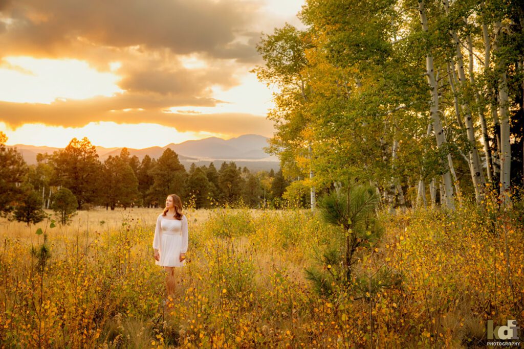 High school senior photos showing girl with blond hair, in white dress among the colorful fall aspen trees, with a dramatic sunset sky, portrait photography in Flagstaff AZ by senior photographer KDI Photography