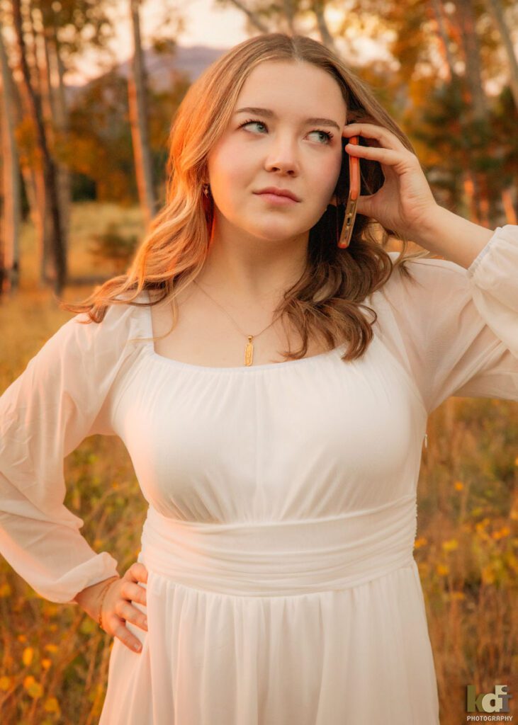 A color photograph of a high school senior photo showing girl with blond hair, in white dress among the fall aspen trees during sunset, talking on a cell phone, portrait photography in Flagstaff AZ by senior photographer KDI Photography