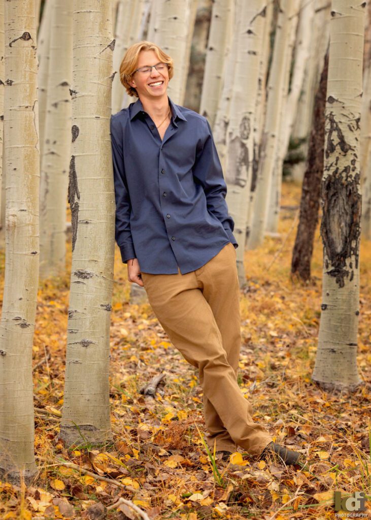 Color portrait a rock climber, senior photos of a senior boy laughing, wearing a blue button down shirt and khaki pants, standing the colorful fall aspens in the mountains of Flagstaff, by family portrait artist, KDI Photography
