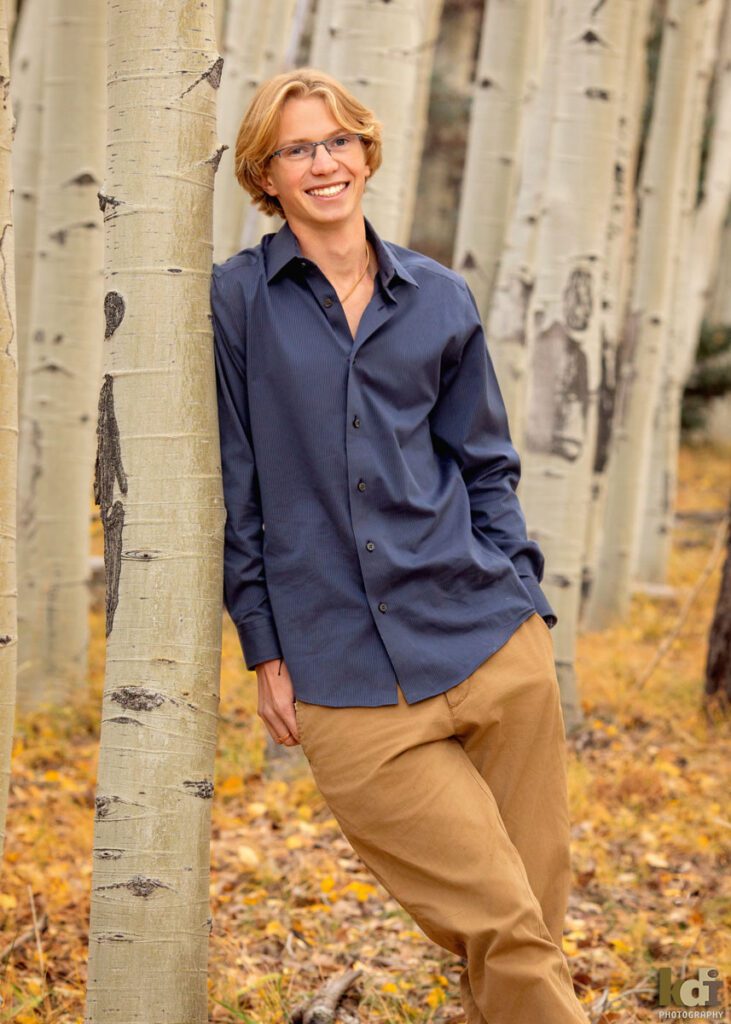 Color portrait a rock climber, senior photos of a senior boy smiling, wearing a blue button down shirt and khaki pants, standing the colorful fall aspens in the mountains of Flagstaff, by family portrait artist, KDI Photography