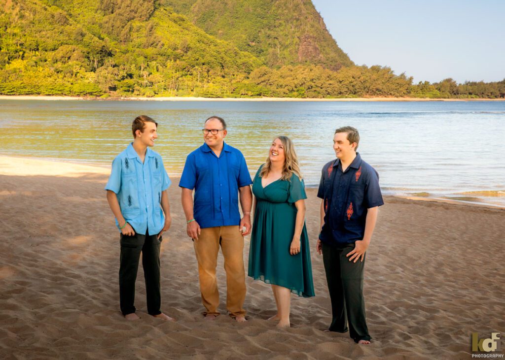 Family of four, mother, father, and two teen boys, laughing and talking  on a beach in Hawaii with mountains and clear blue water in the background, location family photos, by Flagstaff Photographer KDI Photography
