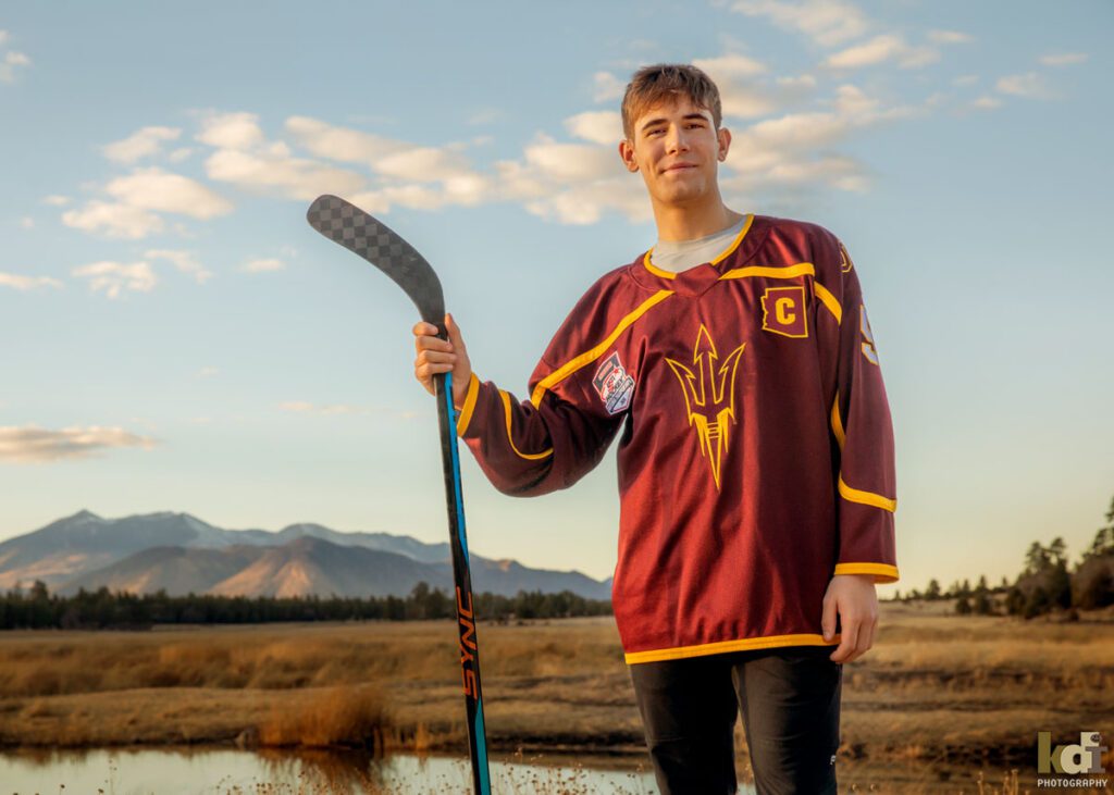 A high school hockey player in a red ASU jersey holds his hockey stick a pond, near the San Francisco Peaks, by Flagstaff photographers, KDI Photography
