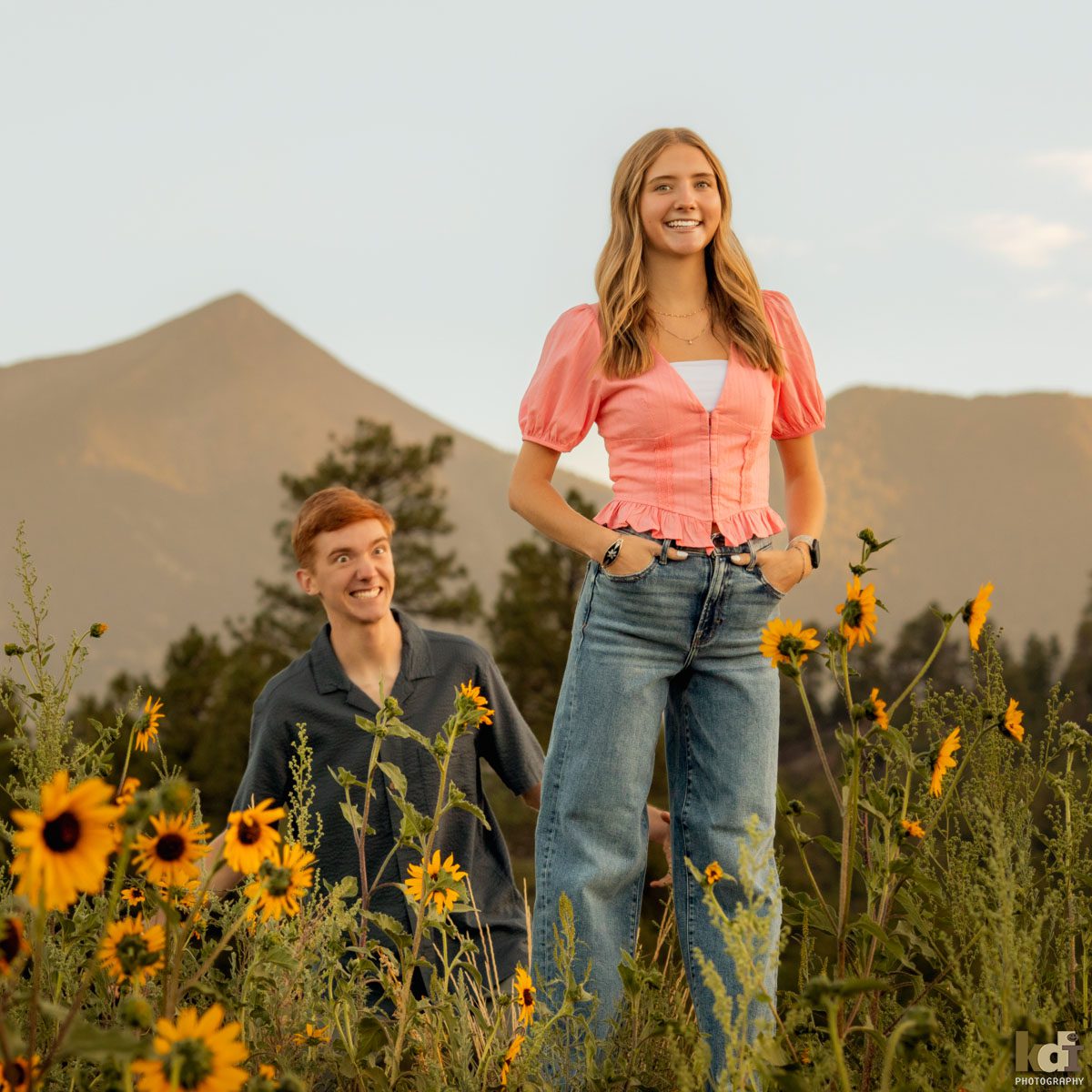 Family photo of redheaded boy in a grey top, sneaking up behind his sister to scare her, in the yellow summer sunflowers, with the San Francisco Peaks in the background, location family portraits by Flagstaff photographer, KDI Photography. 