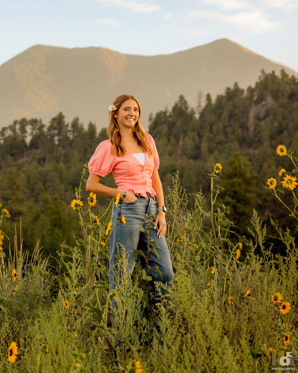 Senior photo of blonde girl in a pink top and jeans, in the wildflowers with the San Francisco Peaks in the background, location senior portraits by Flagstaff photographer, KDI Photography. 