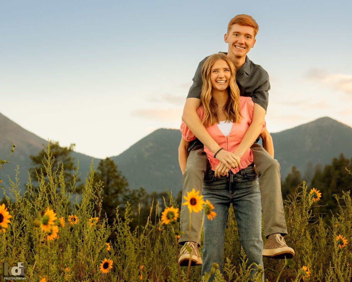 Family photo of blonde girl in a pink top and jeans, giving her brother a piggy back ride, in the yellow wildflowers with the San Francisco Peaks in the background, location family portraits by Flagstaff photographer, KDI Photography. 