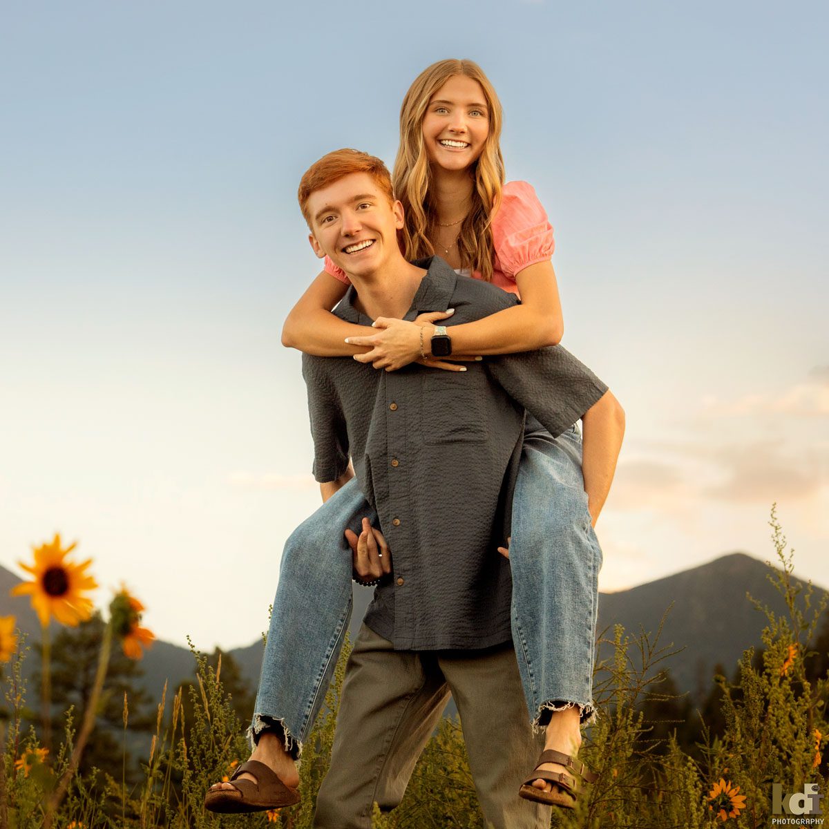 Family photo of redheaded boy in a grey top, giving his sister, a blonde girl in a pink top and jeans, a piggy back ride, in the yellow wildflowers with the San Francisco Peaks in the background, location family portraits by Flagstaff photographer, KDI Photography. 