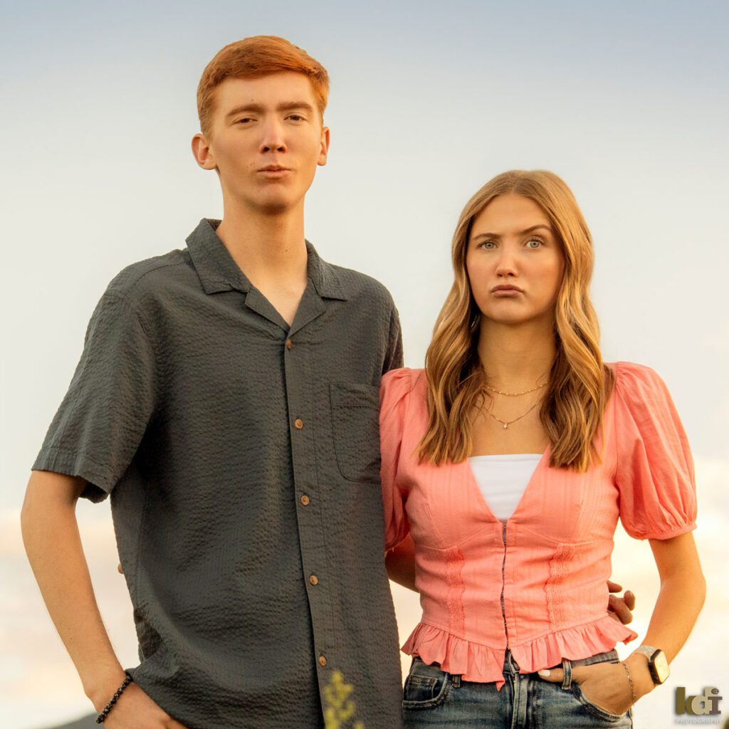 Family photo of brother and sister, making funny faces, each lifting one eyebrow, redheaded boy in a grey top, girl in a pink top and jeans, in the yellow summer sunflowers, with the San Francisco Peaks in the background, location family portraits by Flagstaff photographer, KDI Photography. 