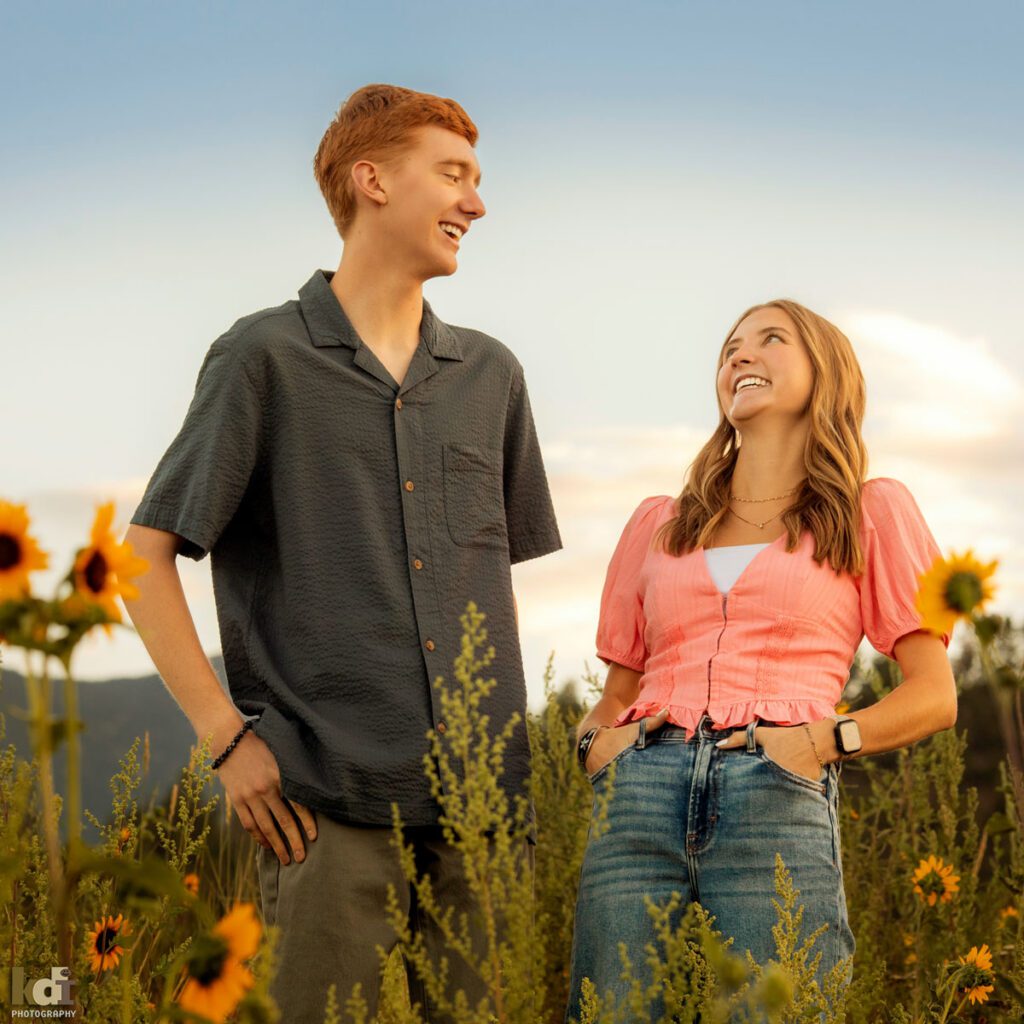 Family photo of brother and sister, facing each other, laughing, redheaded boy in a grey top, girl in a pink top and jeans, in the yellow summer sunflowers, with the San Francisco Peaks in the background, location family portraits by Flagstaff photographer, KDI Photography. 