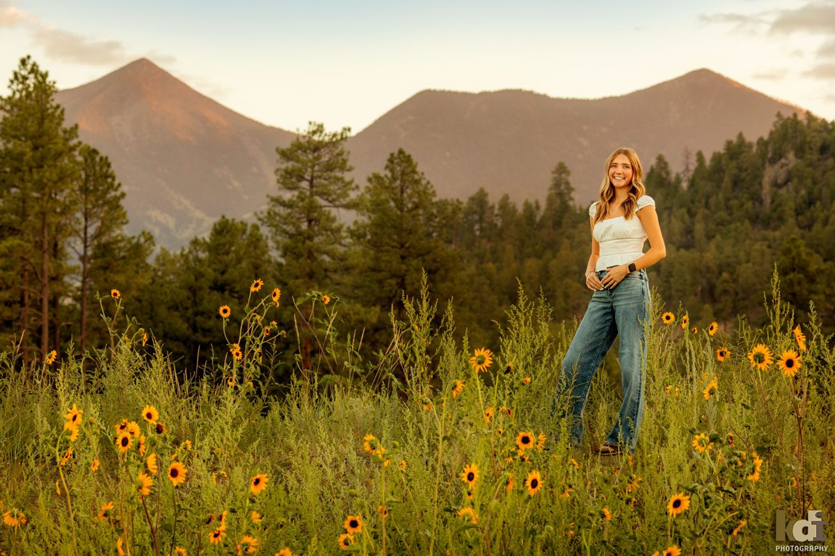 Senior photo of blonde girl in a white top with jeans, in the wildflowers with the San Francisco Peaks in the background, location senior portraits by Flagstaff photographer, KDI Photography. 