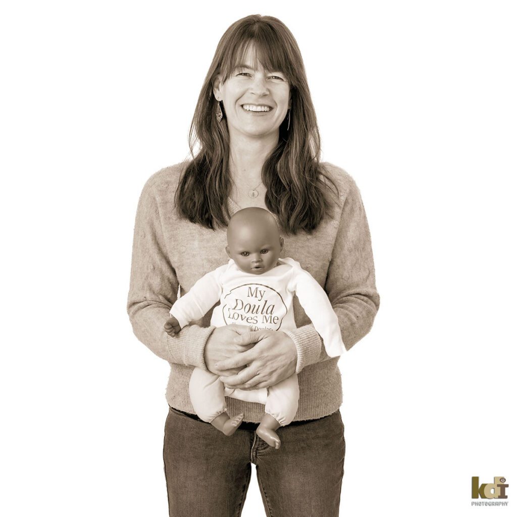 Black and white portrait of a professional doula, holding a baby doll wearing a onsie that says My Doula Loves Me, photography by Flagstaff portrait artist KDI Photography