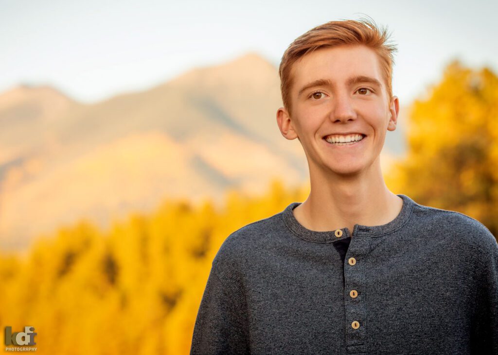Color portrait of redheaded boy in a blue grey shirt, senior in high school, laughing, with the San Francisco Peaks and Ponderosa trees in the background, location family portraits by Flagstaff photographer, KDI Photography. 
