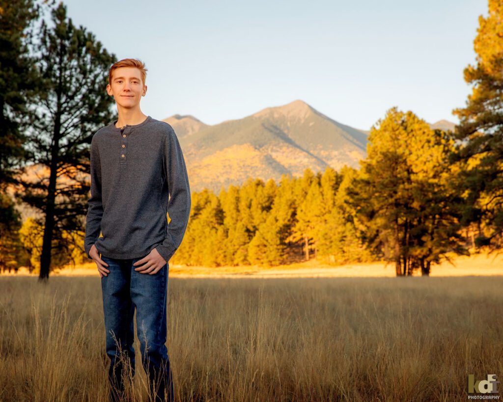 Color portrait of redheaded boy in a grey blue shirt and jeans, senior in high school, with grasses, the San Francisco Peaks and Ponderosa trees in the background, location family portraits by Flagstaff photographer, KDI Photography.