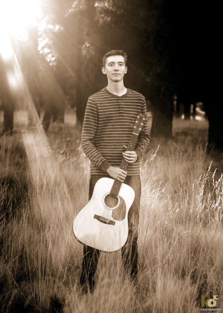 Black and white portrait of senior in high school as he holds his guitar in the forest, with grasses as the sun flared in behind him with Ponderosa trees in the background, location family portraits by Flagstaff photographer, KDI Photography. 