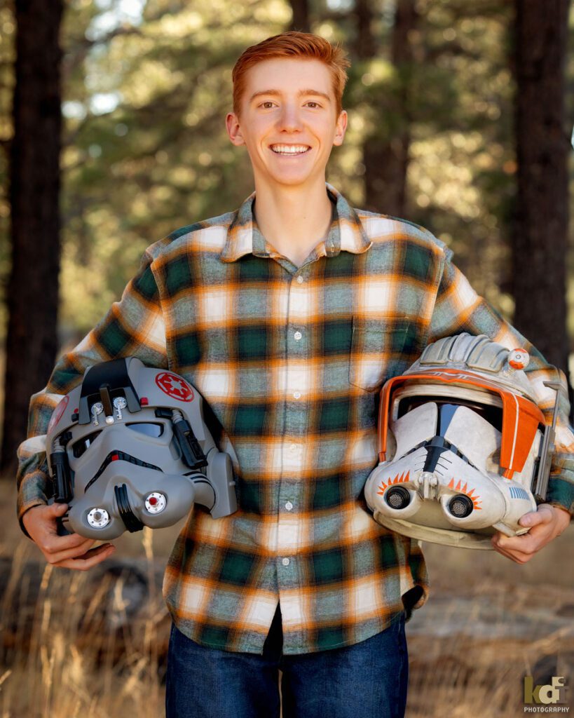 Color portrait of redheaded boy in a red plaid shirt, senior in high school, holding two Star Wars helmets he made with 3D  printing, with grasses and Ponderosa trees in the background, location family portraits by Flagstaff photographer, KDI Photography.