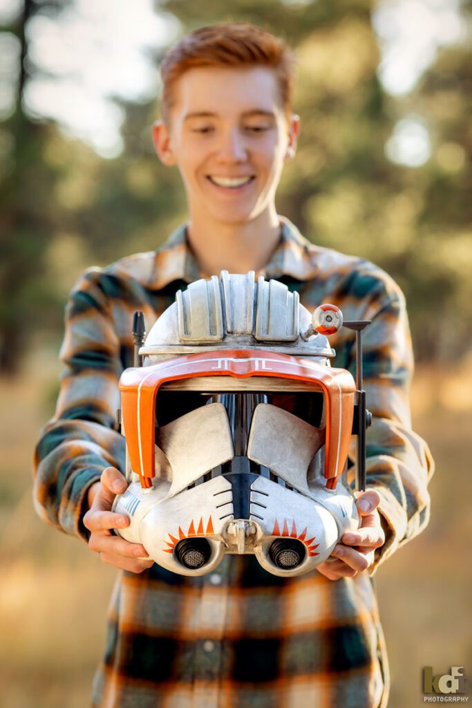 Color portrait of redheaded boy in a red plaid shirt, senior in high school, holding a Star Wars helmet he made with 3D printing, with grasses and Ponderosa trees in the background, location family portraits by Flagstaff photographer, KDI Photography.
