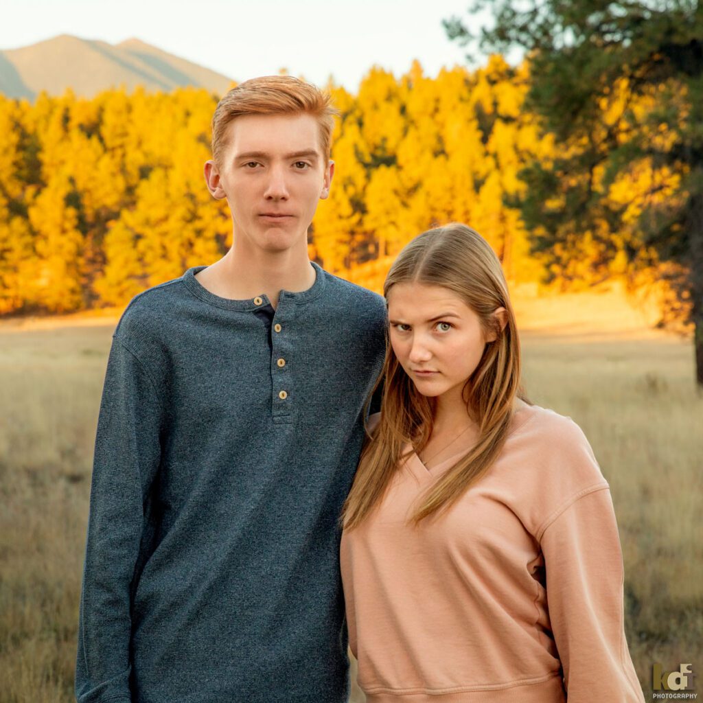 Family photo of brother and sister, making funny faces, each lifting one eyebrow, redheaded boy in a grey top, with Ponderosa trees and the San Francisco Peaks in the background, location family portraits by Flagstaff photographer, KDI Photography. 