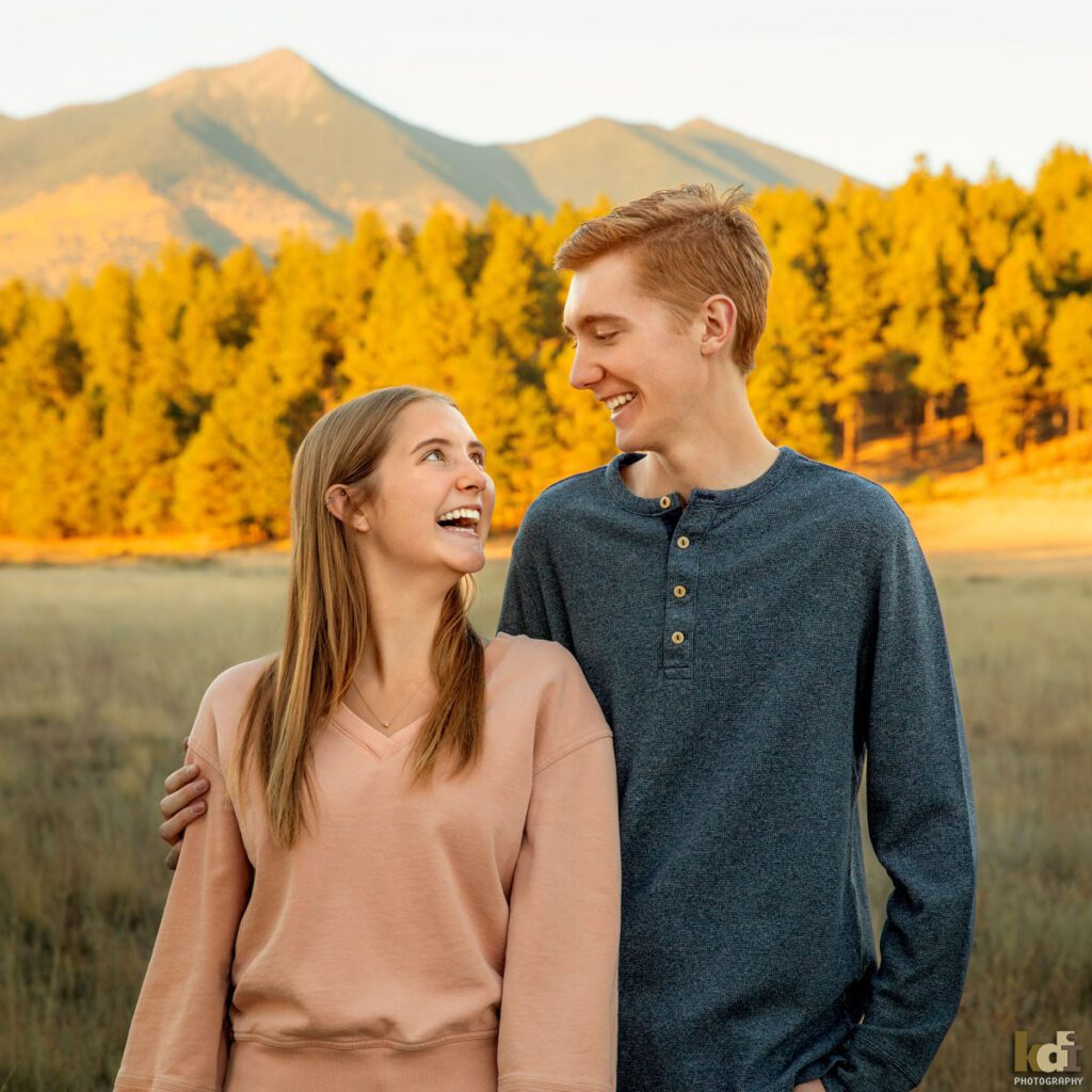 Family photo of brother and sister, laughing as the brother gives his sister a piggy back ride, redheaded boy in a grey top, with Ponderosa trees and the San Francisco Peaks in the background, location family portraits by Flagstaff photographer, KDI Photography. 