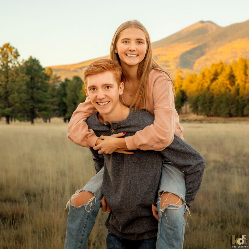 Family photo of brother and sister, laughing as the brother gives his sister a piggy back ride, redheaded boy in a grey top, with Ponderosa trees and the San Francisco Peaks in the background, location family portraits by Flagstaff photographer, KDI Photography. 