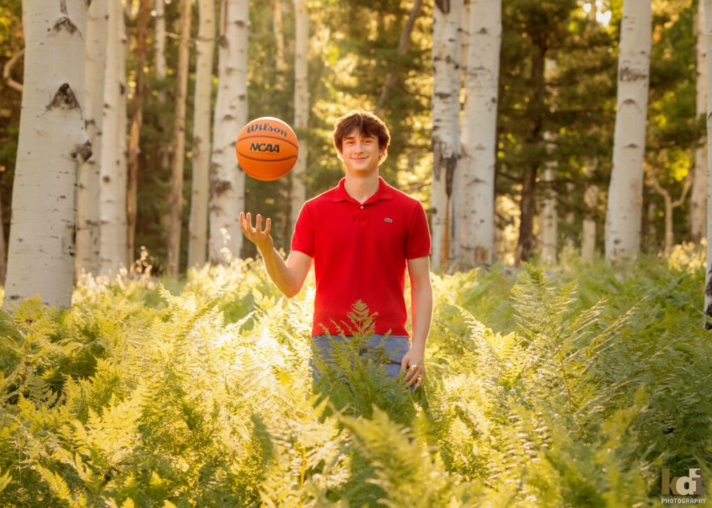 High school senior photos showing boy with brown hair, wearing red shirt, tossing basketball among the summer ferns and aspen trees, portrait photography in Flagstaff AZ by senior photographer KDI Photography