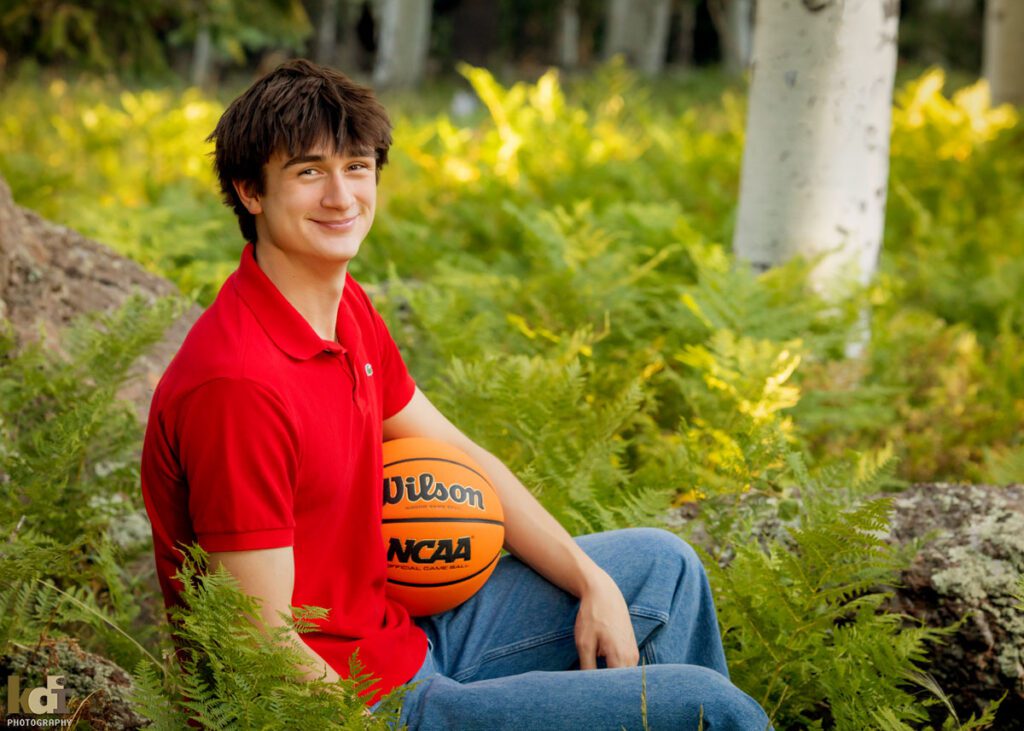 High school senior photos showing boy with brown hair, wearing red shirt, smiling off camera among the summer ferns and aspen trees, portrait photography in Flagstaff AZ by senior photographer KDI Photography
