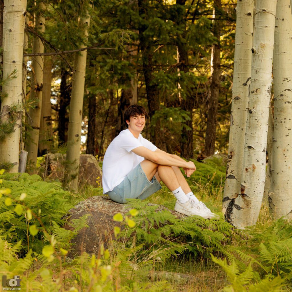 High school senior photos showing boy with brown hair, wearing white t-shirt and jorts, jean shorts, laughing at the camera among the summer ferns and aspen trees, portrait photography in Flagstaff AZ by senior photographer KDI Photography