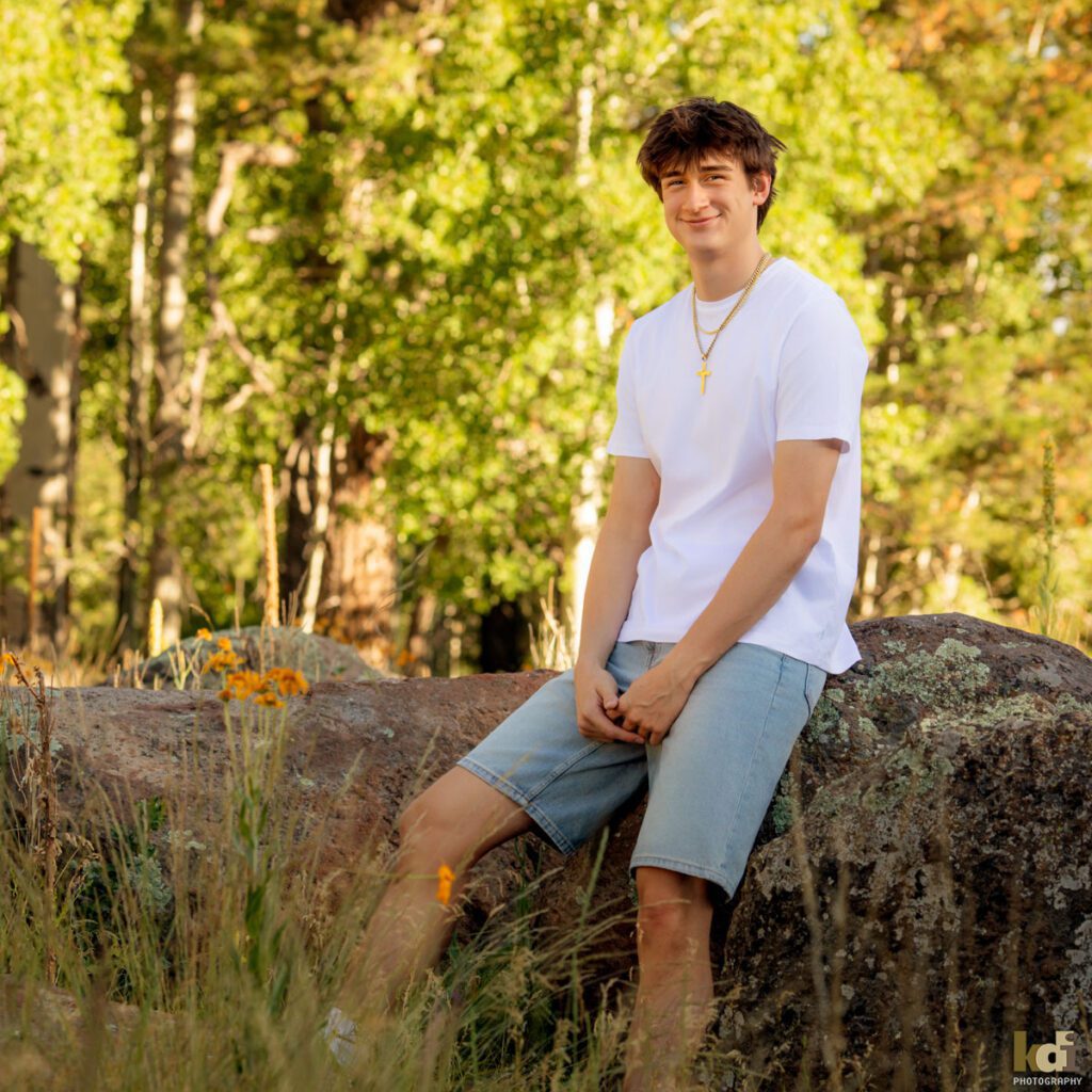 High school senior photos showing boy with brown hair, wearing white t-shirt and jorts, jean shorts, smiling at the camera among the summer ferns and aspen trees, portrait photography in Flagstaff AZ by senior photographer KDI Photography
