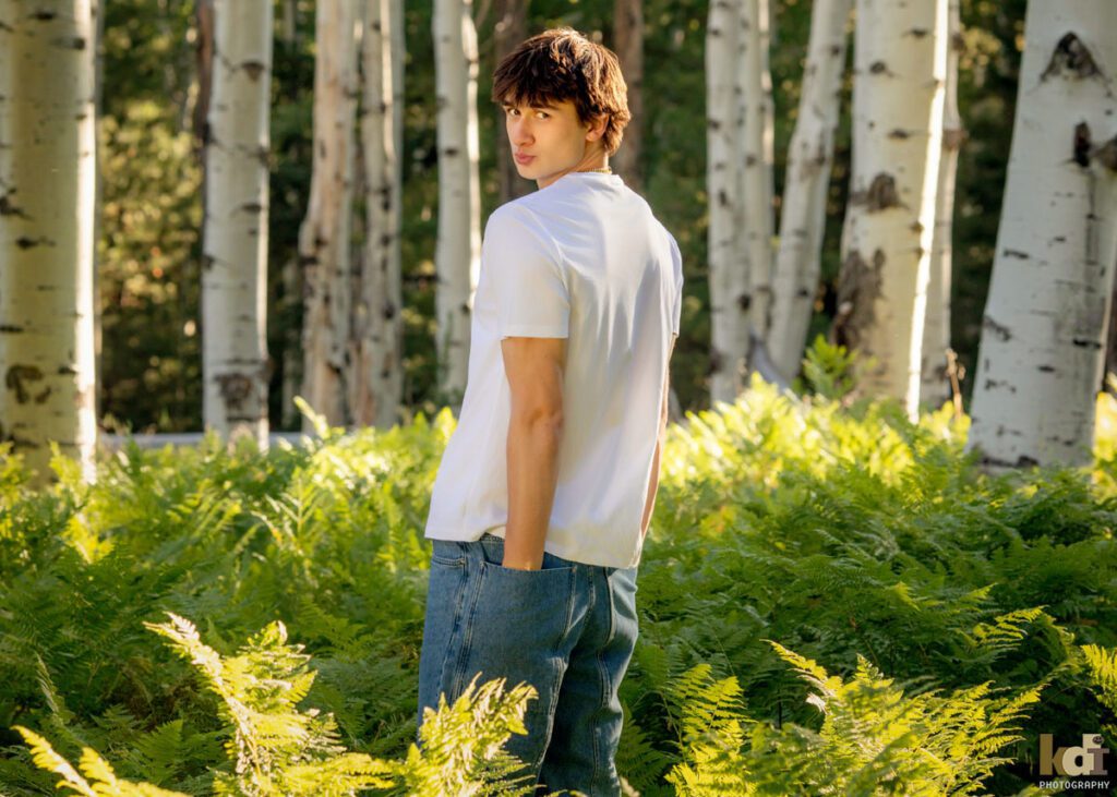 High school senior photos showing boy with brown hair, wearing white shirt, making a face at the camera among the summer ferns and aspen trees, portrait photography in Flagstaff AZ by senior photographer KDI Photography