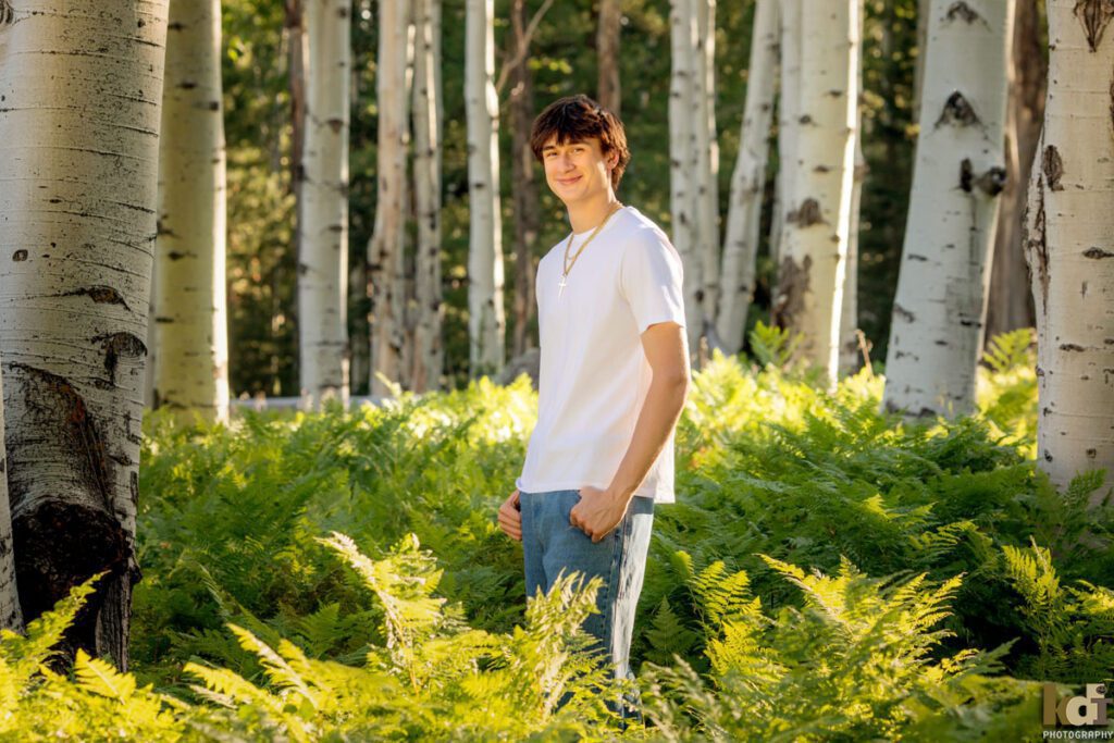 High school senior photos showing boy with brown hair, wearing white shirt, smiling at the camera among the summer ferns and aspen trees, portrait photography in Flagstaff AZ by senior photographer KDI Photography